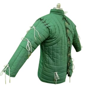 May include: Green padded gambeson with a high collar and long sleeves. The quilted jacket features vertical stitching and is laced with white and black cords. The sleeves are tied with white cords. This medieval garment is designed for historical reenactment or costume.