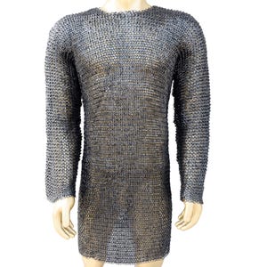 Puede incluir: Una cota de malla de manga larga, exhibida en un maniquí. La camisa está hecha de pequeños anillos de metal entrelazados, creando una prenda protectora y flexible. El color es gris plateado apagado, con una apariencia ligeramente envejecida.