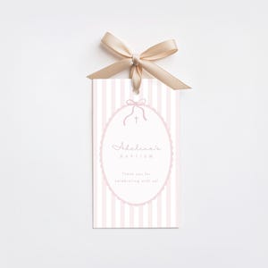 May include: A baptism gift tag with a pink and white striped background. The tag has an oval frame with the text "Adeline's Baptism" and a small cross. A beige ribbon is tied at the top.