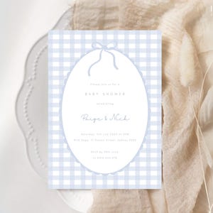 Pu&ograve; includere: Un invito per un baby shower con uno sfondo a quadretti vichy azzurri. L'invito presenta una cornice ovale con un bordo smerlato e un fiocco in alto. Il testo recita "Baby Shower" e i nomi "Paige & Nick".