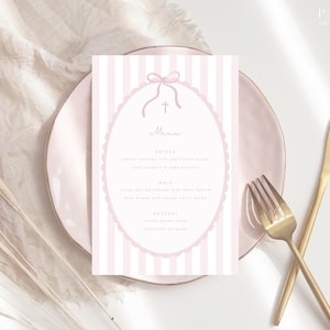May include: A pink and white striped menu card with an oval frame, bow, and cross design, placed on a pink plate with gold cutlery. The menu includes text for a meal. The background includes a white cloth and dried plants.