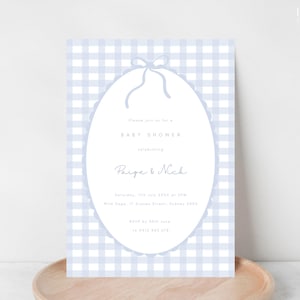 Pu&ograve; includere: Invito per baby shower con sfondo a quadretti vichy azzurri. L'invito presenta una cornice ovale con bordo smerlato e fiocco. Il testo include "Baby Shower" e i nomi "Paige & Nick".
