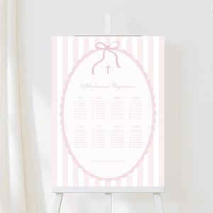 May include: A pink and white striped sign on an easel. The sign features a pink oval frame with a bow and cross, and the text "Adelena's Baptism". Below the text are rows of names. The sign is displayed in front of a white curtain.