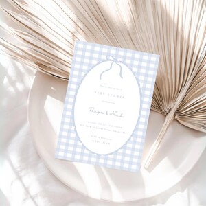 Pu&ograve; includere: Un invito per un baby shower con uno sfondo a motivo gingham azzurro. L'invito presenta un design ovale con un fiocco in alto e le parole "Baby Shower" e i nomi "Paige & Nick". L'invito &egrave; su un piatto bianco con una foglia di palma secca.
