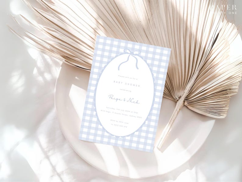 Pu&ograve; includere: Un invito per un baby shower con uno sfondo a motivo gingham azzurro. L'invito presenta un design ovale con un fiocco in alto e le parole "Baby Shower" e i nomi "Paige & Nick". L'invito &egrave; su un piatto bianco con una foglia di palma secca.
