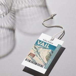 Puede incluir: Una etiqueta blanca de pase de esquí con la palabra "VAIL" en azul. La etiqueta presenta una ilustración de montaña y el texto "1 DAY SKI PASS". Una cuerda está unida a la parte superior de la etiqueta.