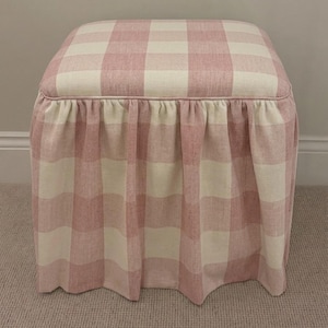 May include: A square ottoman upholstered in a light pink and cream buffalo check fabric. The seat features a piped edge, and the base is finished with a gathered, floor-length skirt that drapes evenly around all four sides of the furniture piece.