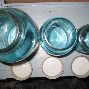 May include: Four blue glass jars with white lids. The jars are different sizes and are arranged in a row.