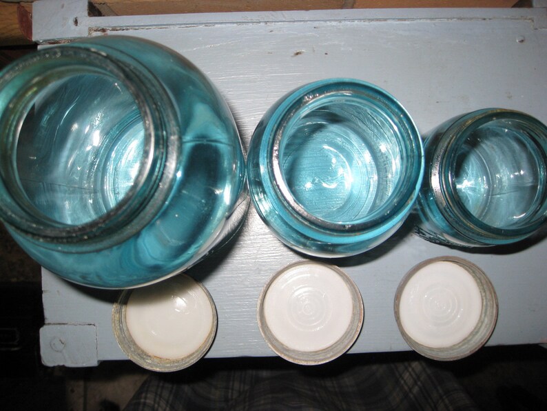 May include: Four blue glass jars with white lids. The jars are different sizes and are arranged in a row.