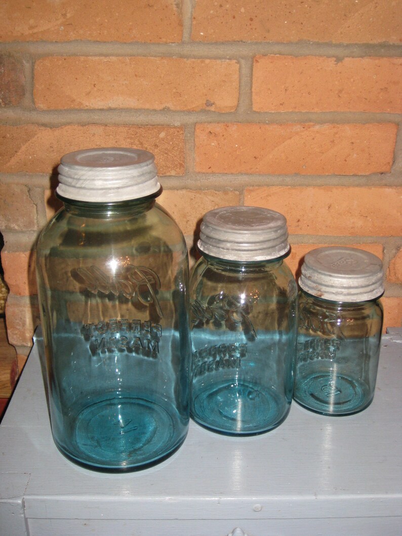 May include: Three blue glass jars with silver lids. The jars are different sizes and have a vintage look. The jars are on a gray surface.
