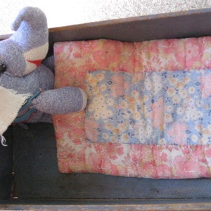 May include: A blue and white sock monkey toy lying in a wooden bed with a pink and blue floral patterned quilt.