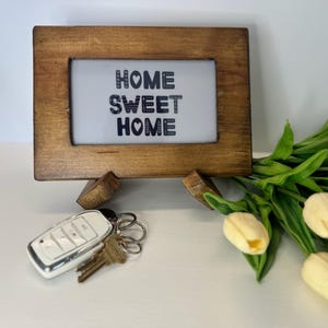以下が含まれることがあります： 白い背景に「HOME SWEET HOME」という黒い文字が書かれた小さな木製のフレーム。フレームは小さな木製のスタンドの上に置かれています。白い車のキーとキーのセットが手前にあります。背景には黄色いチューリップのブーケがあります。