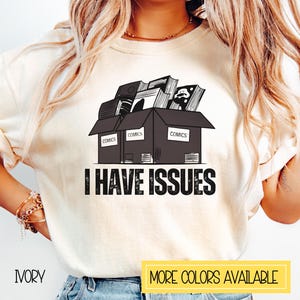 May include: A white t-shirt with a black and white graphic of a box overflowing with comic books. The box has the word "COMICS" printed on each side. The text "I HAVE ISSUES" is printed below the box.