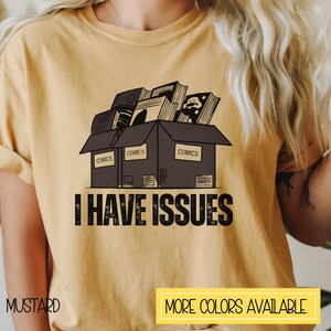 May include: A mustard yellow t-shirt with a graphic of a box overflowing with comic books. The box is labeled "COMICS" on each side. The text "I HAVE ISSUES" is printed below the box.