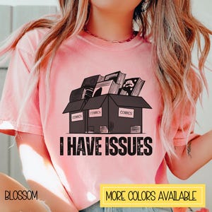May include: Pink t-shirt with a graphic of three boxes stacked with comic books inside. The boxes are labeled "COMICS". The text "I HAVE ISSUES" is printed below the boxes.