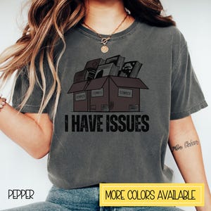May include: A gray t-shirt with a graphic of three boxes stacked on top of each other. The boxes are labeled "COMICS" and are filled with comic books. The text "I HAVE ISSUES" is printed below the boxes.