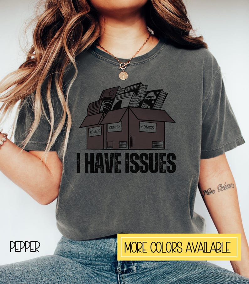 May include: A gray t-shirt with a graphic of three boxes stacked on top of each other. The boxes are labeled "COMICS" and are filled with comic books. The text "I HAVE ISSUES" is printed below the boxes.