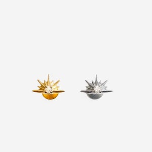 May include: A pair of earrings featuring a starburst design with a small diamond in the center. The earrings are available in gold and silver.