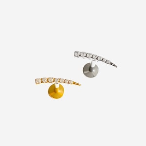 May include: Two curved stud earrings, one gold-toned and one silver-toned, each adorned with a row of small, clear gemstones. The earrings are set against a white background. The brand name "HYANGMI GU Jewelry" is at the bottom.