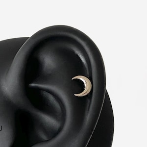 May include: A black ear model with three silver earrings. The earrings are a crescent moon, a star, and a small rectangle with a chain hanging from it.