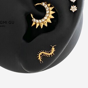 May include: A display of gold earrings on a black ear-shaped form. The earrings include a sunburst hoop, a star, a square, a flower, and a centipede. The text "HYANGMI GU Jewelry" is visible on the form.