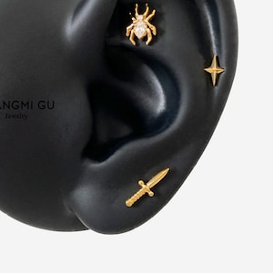May include: A black ear model with three gold earrings: a spider with a white stone, a star, and a sword.