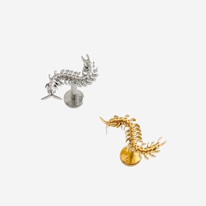 May include: Two centipede-shaped earrings, one silver-toned and one gold-toned, are displayed against a white background. The earrings have a detailed, segmented design and are mounted on posts. The brand name "HYANGMI GU Jewelry" is at the bottom.