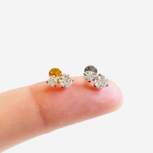 May include: Two stud earrings, each featuring two round, clear, faceted stones set in a prong setting. One earring has a gold-tone metal base, while the other has a silver-tone metal base. Both pieces are designed with a flat back post.