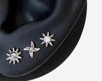May include: Three silver-toned stud earrings arranged in a row. The first is a sunburst shape with a central dome, the second is a four-pointed star with a small clear stone, and the third is a multi-pointed star with a central clear stone.