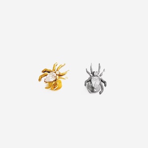 May include: Two spider-shaped stud earrings. One earring is gold-toned with a pear-shaped clear stone on the abdomen and a small round stone on the head. The other earring is dark silver-toned with a matching pear-shaped clear stone and round stone.