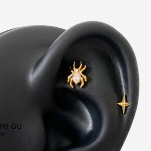 May include: A gold spider-shaped earring stud featuring a central marquise-cut clear gemstone body and a four-pointed star-shaped earring stud positioned below it, both displayed on a black ear-shaped model for jewelry presentation.