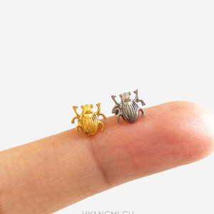 May include: Two small beetle-shaped stud earrings, one in a polished gold tone and the other in a silver tone. Each earring features a detailed, textured shell with visible legs and antennae, designed as a pair of insect-themed jewellery pieces.