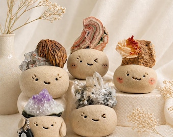 Puede incluir: Cinco pequeñas macetas de cerámica beige, redondas, con caras sonrientes dibujadas a mano. Cada maceta contiene un espécimen natural diferente: un grupo mineral marrón, una rodaja de ágata, una roca texturizada, amatista y cuarzo transparente.