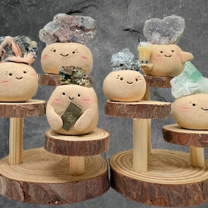 Puede incluir: Una colección de figuras de cerámica beige, caprichosas y sonrientes, cada una adornada con un tipo diferente de cristal o piedra preciosa. Las figuras se exhiben sobre soportes de madera con una estética natural y rústica. Una figura sostiene un billete pequeño.