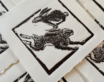 May include: A collection of square art prints featuring a linocut design of two running hares. The hares are depicted in black ink within a black square border on a white paper background. The prints have a textured, handmade appearance.