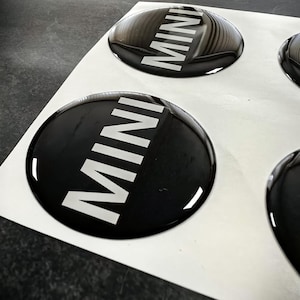 May include: A sheet of black and white MINI logo stickers. The stickers are round with a glossy finish. The word "MINI" is printed in white, bold letters on a black background.