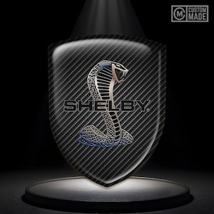 May include: A black and silver Shelby Cobra logo with a carbon fibre background. The logo is in the shape of a shield and features a snake with a raised hood.