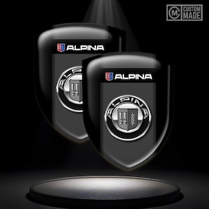 May include: Two black and silver Alpina car emblems with the word "ALPINA" in white letters on a black background. The emblems are on a black surface with a spotlight shining on them. The text "CUSTOM MADE" is in the upper right corner of the image.