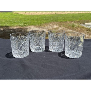 Puede incluir: Cuatro vasos transparentes de cristal tallado con un detallado patrón floral y geométrico. Los vasos son cilíndricos con una base gruesa y están dispuestos sobre una superficie oscura. El diseño de cristal tallado crea un efecto brillante.