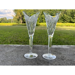 May include: Two clear crystal champagne flutes with detailed etched patterns. The flutes feature a long stem and a wide bowl, ideal for special occasions. The glasses are placed on a grey surface with a green backdrop.
