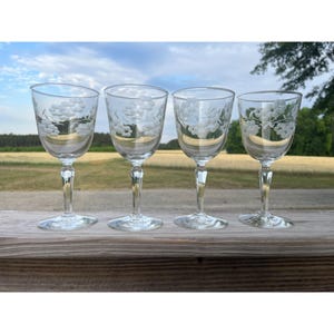 May include: A set of four clear glass wine glasses, each featuring a delicate etched floral pattern around the upper bowl. The glasses have a slender stem and a circular base, showcasing a transparent design with white frosted floral details.