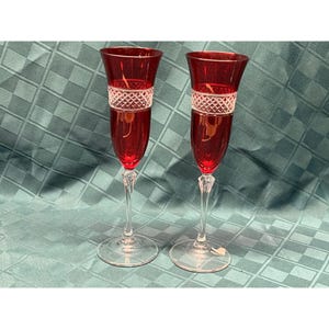 May include: Two red champagne flutes with a clear, diamond-patterned band around the middle. The stems are clear and have a decorative design. The flutes are on a teal tablecloth.