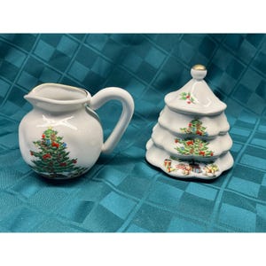 May include: A white ceramic creamer and a matching lidded sugar bowl shaped like a tiered Christmas tree. Both pieces feature colorful illustrations of decorated pine trees, red bows, and various toys, finished with gold trim on the edges.