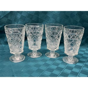 May include: Four clear, vintage-style drinking glasses. Each glass features a diamond-cut pattern and sits on a short, round pedestal base. The glasses are arranged on a teal-colored tablecloth.
