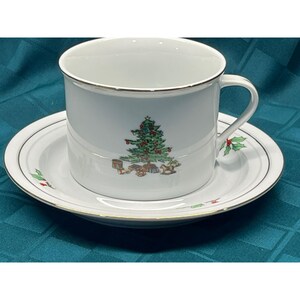 May include: A white ceramic teacup and saucer set featuring a gold rim and a central illustration of a decorated Christmas tree with gifts underneath. The saucer also displays small green holly leaf accents with red berries along the edge.