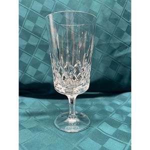 May include: A clear glass goblet featuring a tall, cylindrical bowl with a faceted diamond-cut pattern on the lower half. The stem is short and slender, connecting to a circular, flat base. The glass is transparent with reflective surfaces.
