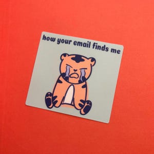 May include: A sticker with a cartoon tiger illustration. The tiger is orange with black stripes and is crying. The text "how your email finds me" is above the tiger on a light blue background, set against a red background.