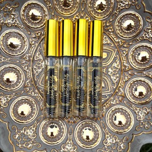 Può includere: Quattro flaconi di vetro trasparente di "Eyelash Serum" con tappi dorati ed etichette nere, disposti su un piatto decorativo grigio e oro. Il piatto presenta un design ornato in rilievo con motivi circolari e dettagli dorati. Il testo "Nails By Milly" è visibile.