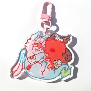 May include: A clear acrylic keychain featuring a cartoon illustration of two creatures embracing. One is pink and blue with bunny-like features, the other is red with brown wings. The keychain has a pink clasp.