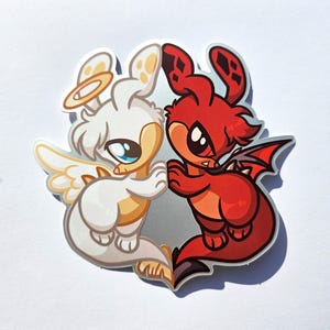May include: A sticker featuring two cartoon characters: a white angel with a halo and wings, and a red devil with horns and a tail. The angel and devil are hugging each other. The sticker has a glossy finish.